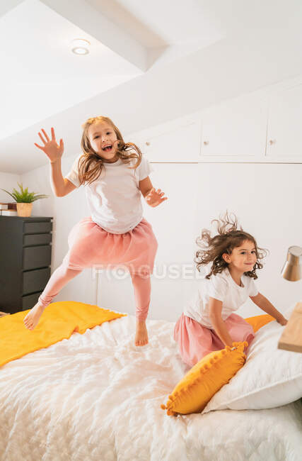 Full body delighted little sisters in casual skirts jumping happily on soft cozy bed while having fun together in light bedroom — Stock Photo
