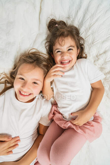 Возбужденные маленькие сестры в повседневной одежде, лежащие на удобной кровати вместе, глядя в камеру — стоковое фото