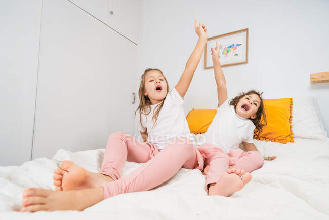Возбужденные маленькие сестры в повседневной одежде лежат на удобной кровати и поднимают руки вместе, глядя в камеру — стоковое фото