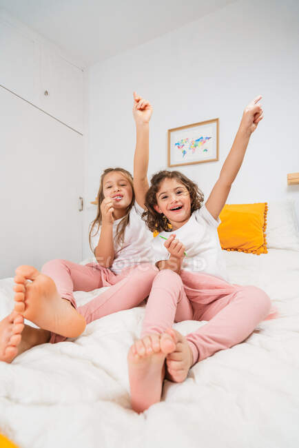 Захоплені маленькі сестри в повсякденному одязі лежать на зручному ліжку і піднімають руки щасливо, співаючи улюблену пісню разом — стокове фото