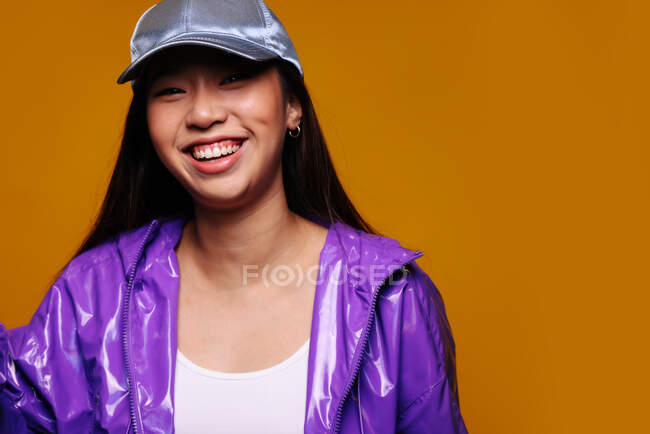 Retrato de la joven asiática feliz. Lleva una chaqueta púrpura y una gorra gris y mira a la cámara sonriendo sobre un fondo amarillo. - foto de stock