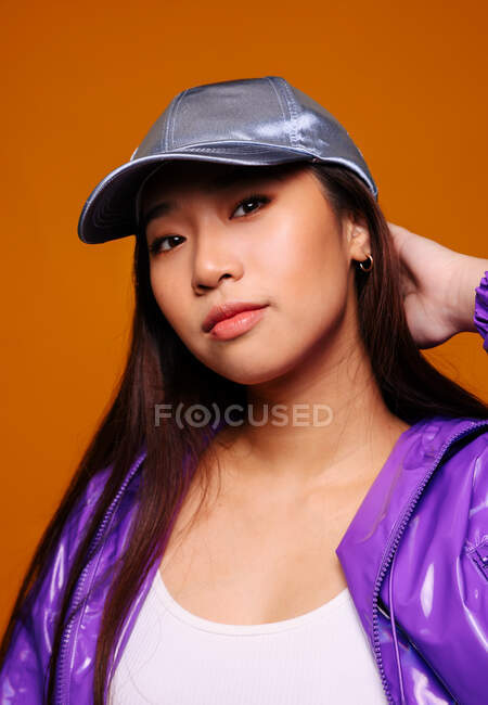 Retrato de mulher jovem asiática com expressão séria. Ela usa uma jaqueta roxa e um boné cinza e está olhando para a câmera contra um fundo amarelo enquanto toca a cabeça. — Fotografia de Stock