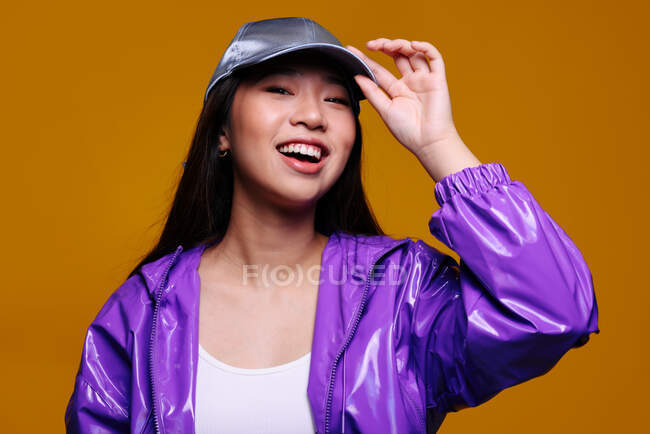 Retrato da jovem asiática feliz. Ela usa uma jaqueta roxa e um boné cinza e está olhando para a câmera sorrindo contra um fundo amarelo — Fotografia de Stock