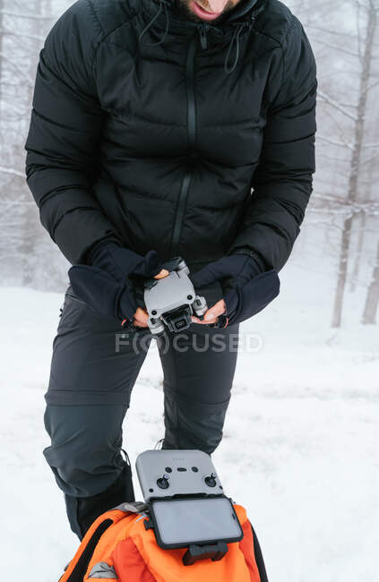 Cultiver jeune homme portant des vêtements chauds noirs tenant drone plié moderne tout en se tenant debout dans les bois verglacés enneigés — Photo de stock
