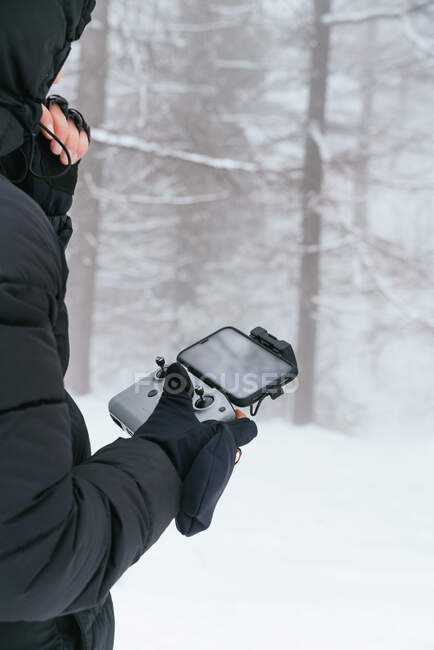 Cropped mâle méconnaissable portant une veste noire chaude avec capuche debout sur un terrain enneigé avec télécommande par drone — Photo de stock