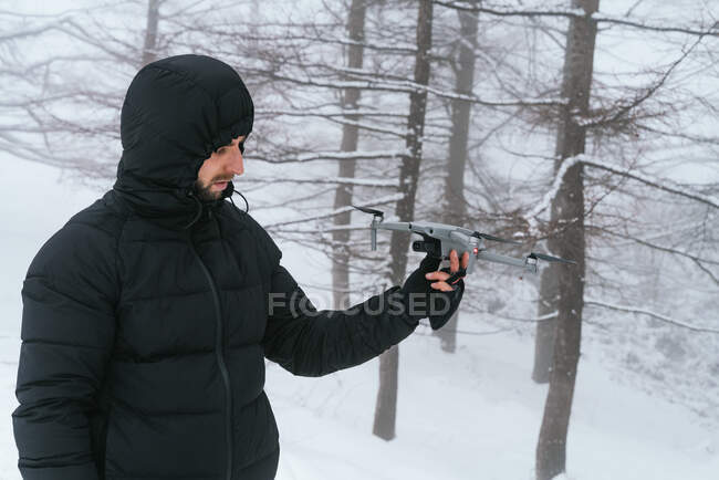 Concentrated young male in warm winter clothes and hood holding contemporary UAV in snowy freezing forest — Stock Photo