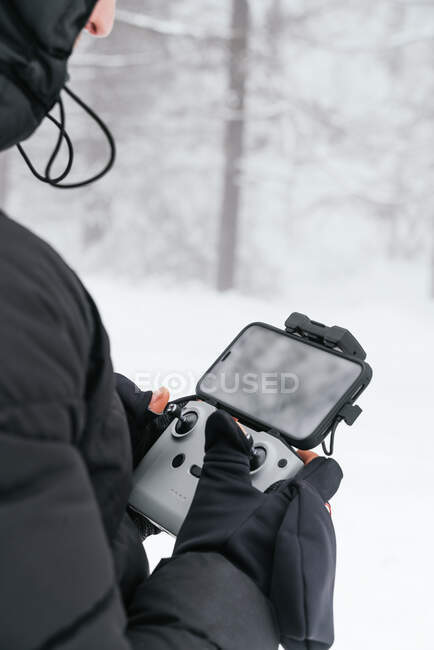Cropped unrecognizable male wearing warm black jacket with hood standing on snowy terrain with drone remote control — Stock Photo