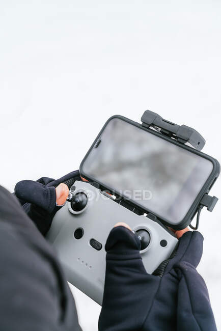 Cropped mâle méconnaissable portant une veste noire chaude avec capuche debout sur un terrain enneigé avec télécommande par drone — Photo de stock