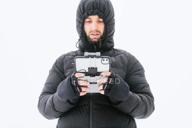 Focused bearded male wearing warm black jacket with hood standing on snowy terrain with drone remote control and looking away — Stock Photo