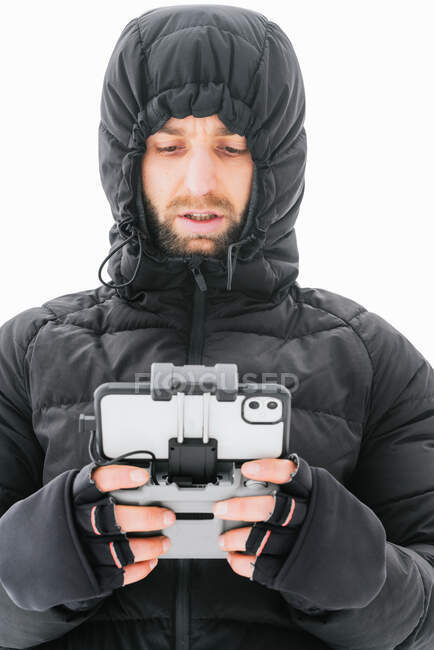 Hombre barbudo enfocado con chaqueta negra cálida con capucha de pie en terreno nevado con control remoto de drones y mirando hacia otro lado - foto de stock