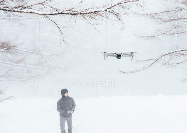 Hombre vistiendo ropa de invierno caliente manteniendo el control remoto de los modernos UAV volando cerca en bosques congelados - foto de stock