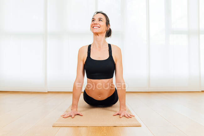 Vue de face femme gaie faisant l'exercice Bhujangasana pendant la séance de yoga à la maison sur tapis de sport — Photo de stock