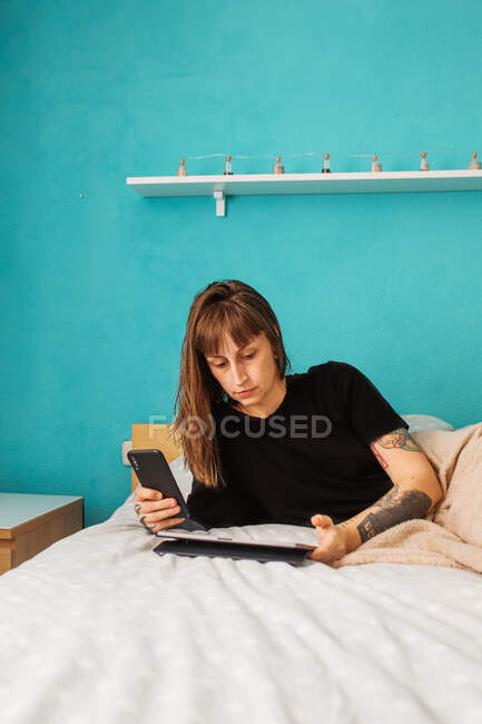 Концентрированная молодая женщина с татуированной рукой просматривает современный планшет и отдыхает на удобной кровати в светлой спальне — стоковое фото