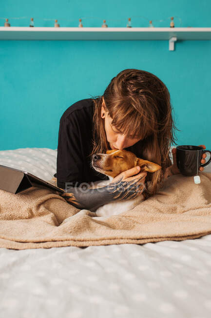 Femme sereine en chemise décontractée avec bras tatoué caressant et embrassant adorable chien de race tout en se reposant ensemble sur un lit confortable avec tablette et tasse de thé — Photo de stock