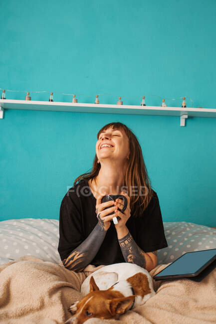 Улыбайтесь женщина в повседневной черной рубашке с татуированными руками наслаждаясь чашкой чая и сидя на уютной кровати с глазами рядом с современной таблеткой и очаровательной спящей собакой — стоковое фото