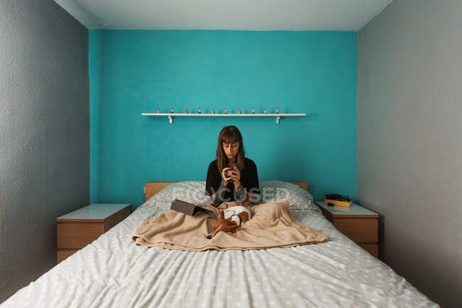 Junge Frau im schwarzen Hemd genießt heißen Tee und schaut sich Videos auf dem Tablet an, während sie sich auf einem bequemen Bett mit einem entzückenden freundlichen Hund ausruht — Stockfoto
