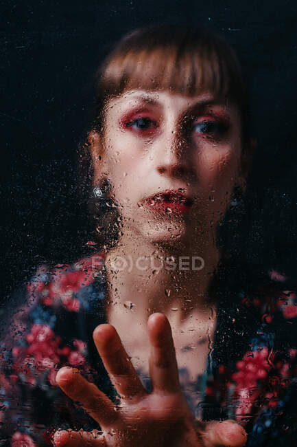 Jeune femme triste en tenue ornementale avec bang regardant caméra derrière verre transparent avec gouttes d'eau — Photo de stock