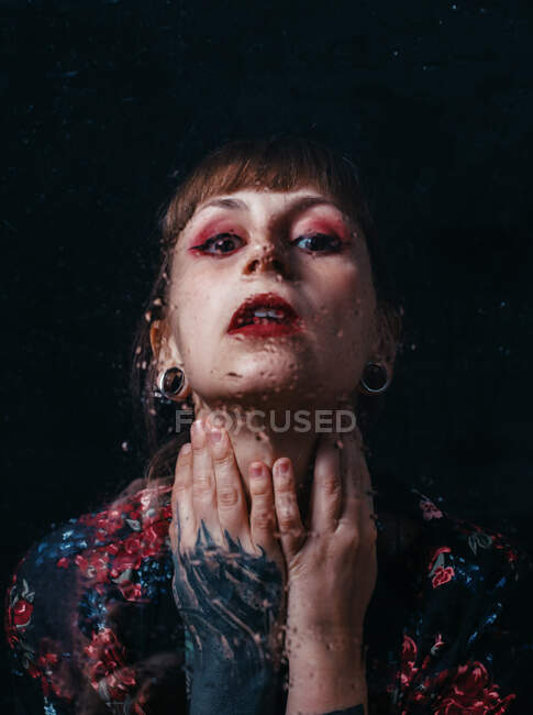 Femme sans émotion avec bras tatouage touchant le cou debout derrière le verre translucide avec des gouttelettes d'eau regardant la caméra — Photo de stock
