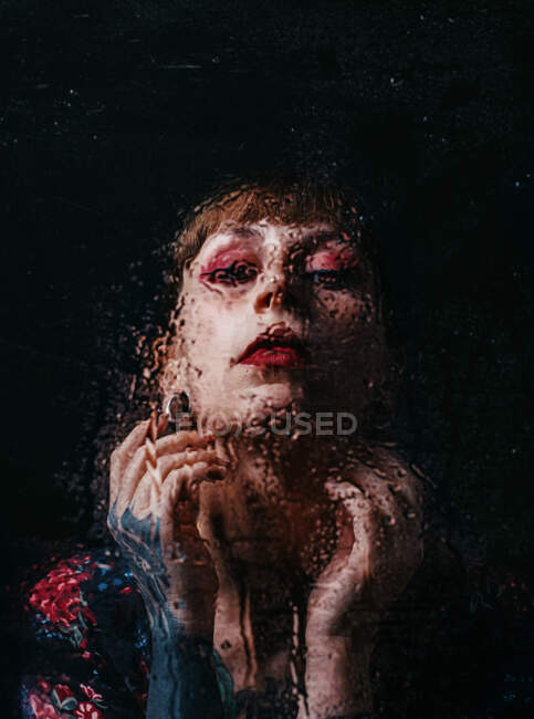 Mujer sin emociones con el brazo tatuaje tocando el cuello de pie detrás de vidrio translúcido con gotas de agua mirando a la cámara - foto de stock