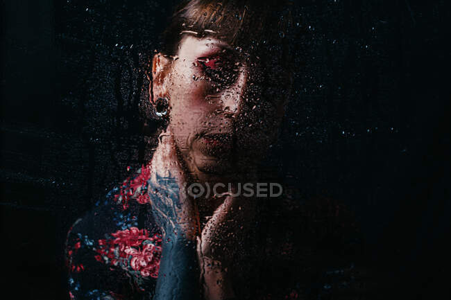 Обрізати засмучена жінка з закритими очима і пофарбована рука торкається шиї за напівпрозорим склом з краплями води — стокове фото
