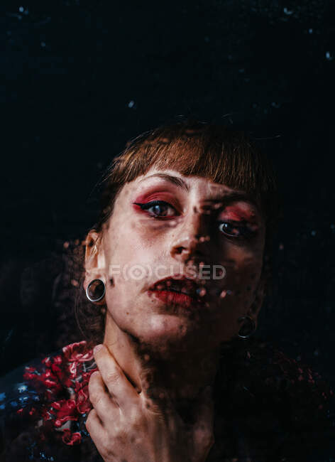 Mujer sin emociones con el brazo tatuaje tocando el cuello de pie detrás de vidrio translúcido con gotas de agua - foto de stock