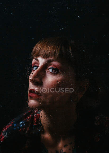 Mujer sin emociones con piercing en el oído de pie detrás de vidrio translúcido con gotitas de agua mirando a la cámara - foto de stock