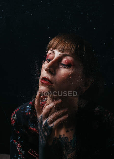 Unemotional female with arm tattoo standing behind translucent glass with water droplets — Stock Photo