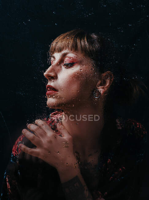 Unemotional female with arm tattoo standing behind translucent glass with water droplets — Stock Photo