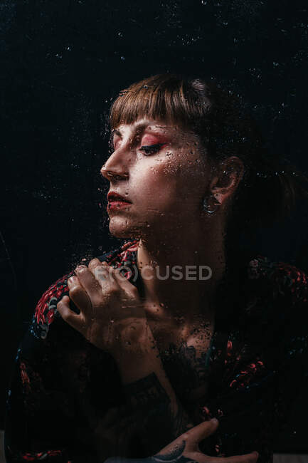 Crop upset female with closed eyes standing behind translucent glass with water droplets — Stock Photo