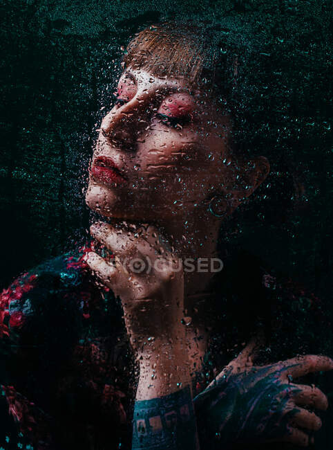 Crop upset female with closed eyes and painted arm touching neck behind translucent glass with water droplets — Stock Photo