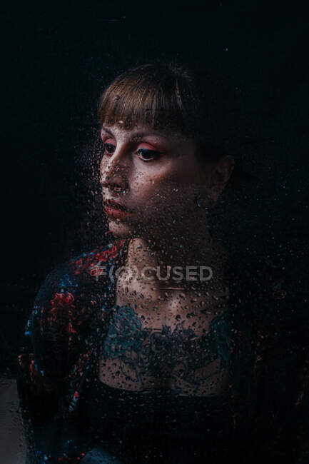 Mujer sin emociones con tatuaje detrás de vidrio translúcido con gotitas de agua mirando hacia otro lado - foto de stock