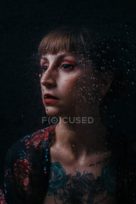 Femme sans émotion avec tatouage debout derrière un verre translucide avec des gouttelettes d'eau regardant loin — Photo de stock
