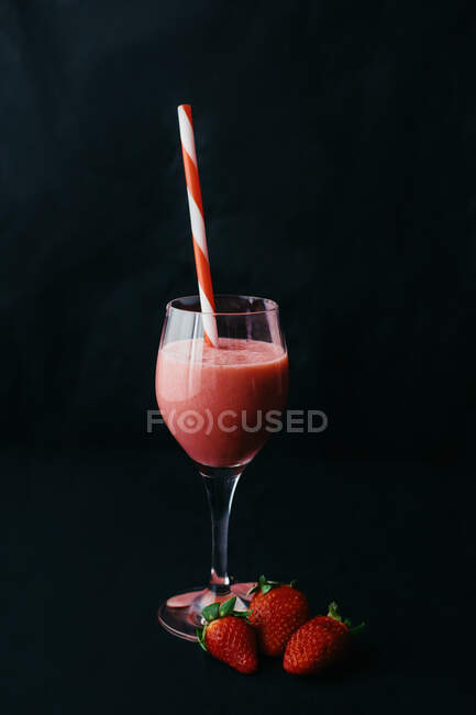 Flow of yummy beverage pouring into transparent glass with striped straw near juicy strawberries on black background — Stock Photo