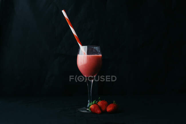 Flusso di bevanda gustosa versando in vetro trasparente con paglia rigata vicino a succose fragole su sfondo nero — Foto stock