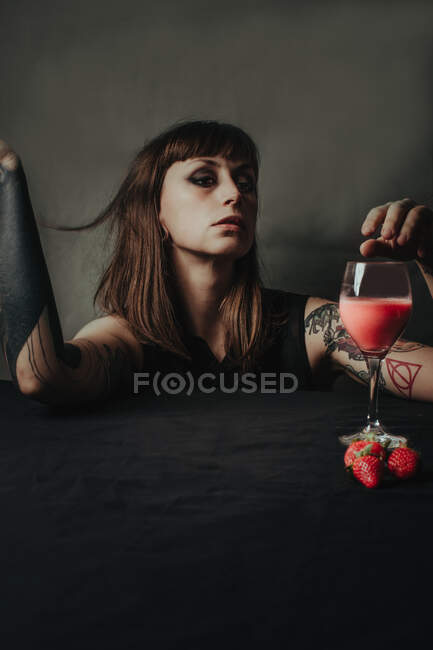 Молодая женщина с темным макияжем и поднятыми руками против прозрачного стакана сладкого освежающего напитка со свежей клубникой — стоковое фото