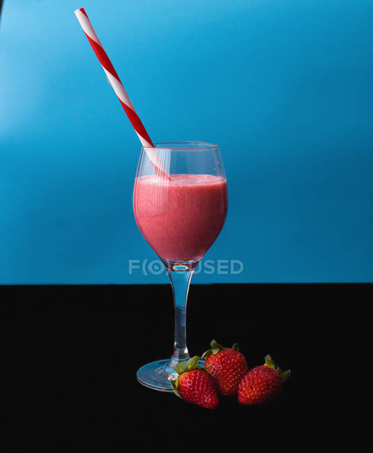 Flow of yummy beverage pouring into transparent glass with striped straw near juicy strawberries on blue background — Stock Photo
