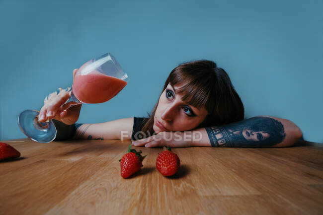 Молодая созерцательная женщина с макияжем и татуировкой, держа стакан клубничного смузи, опираясь рукой на стол на синем фоне — стоковое фото