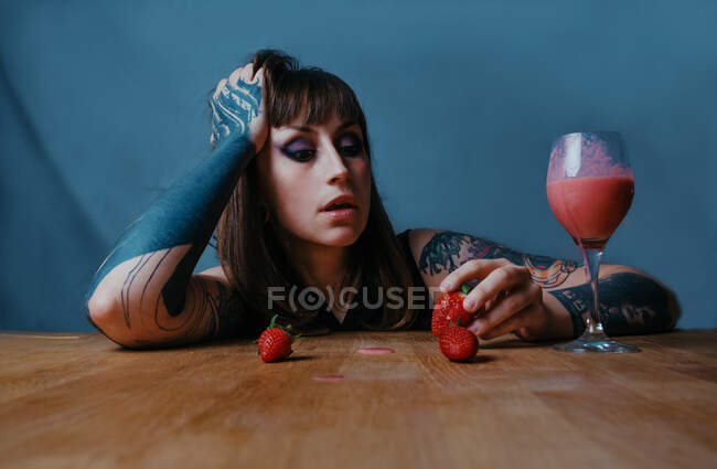 Junge kontemplative Frau mit Make-up und Tätowierung hält Erdbeere, während sie sich mit der Hand auf einen Tisch auf blauem Hintergrund lehnt — Stockfoto