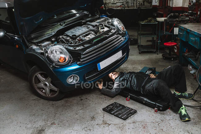 Jovem focado auto serviço homem verificando o veículo com tampa aberta enquanto deitado no tapete na oficina — Fotografia de Stock