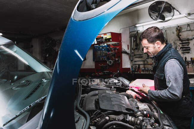 Side view of unshaven maintenance male pouring liquid from canister into radiator in open hood of vehicle in garage — Stock Photo