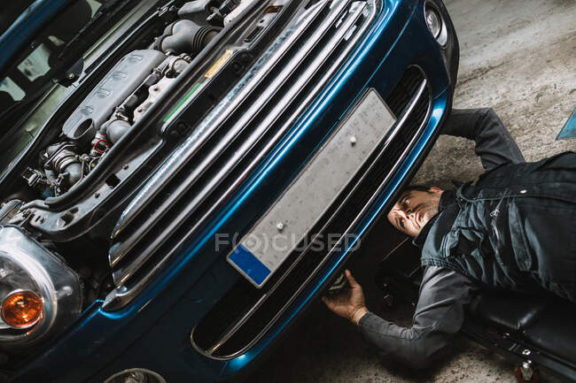 Jovem focado auto serviço homem verificando o veículo com tampa aberta enquanto deitado no tapete na oficina — Fotografia de Stock