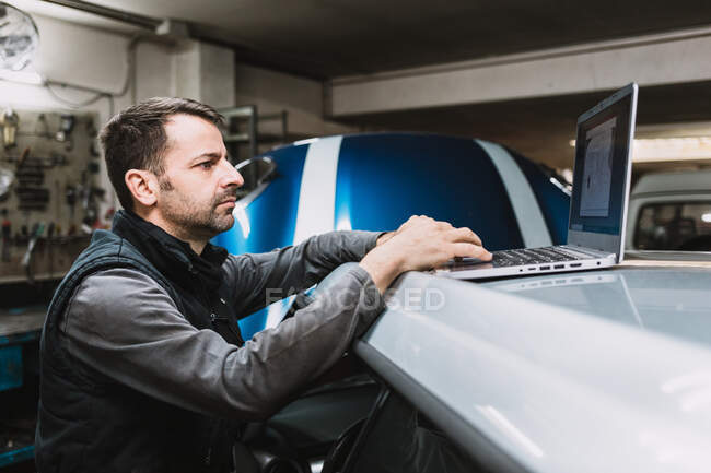 Вид збоку молодого фокусованого бородатого майстра, який дивиться екран нетбука на вершині автомобіля в автомайстерні — стокове фото