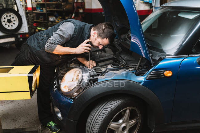 Молодой сфокусированный бородатый ремонтник ремонтирует современный автомобиль против яркого света в мастерской — стоковое фото