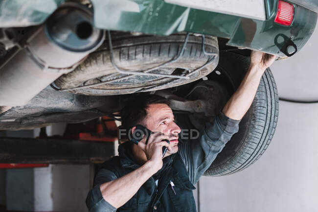 Young focused male auto master speaking on cellphone while checking up rear part of transport and looking up in workshop — Stock Photo