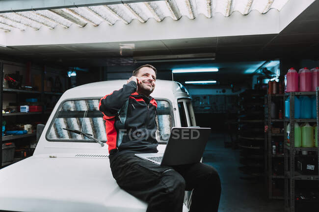 From below of male master with netbook talking on cellphone and sitting on car in garage — Stock Photo