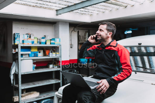 Мужчина мастер с нетбуком разговаривает по мобильному телефону рядом с транспортом в гараже — стоковое фото