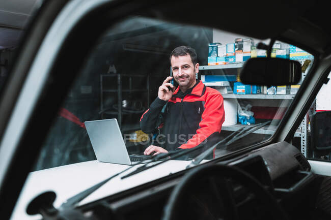 Чоловічий майстер з нетбуком, що розмовляє на мобільному телефоні біля транспорту в гаражі — стокове фото