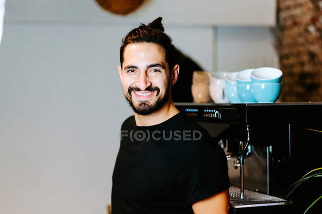 Barista masculino sonriente usando portafilter y preparando café en la cafetera moderna mientras está parado en el mostrador en la cafetería y mirando a la cámara - foto de stock