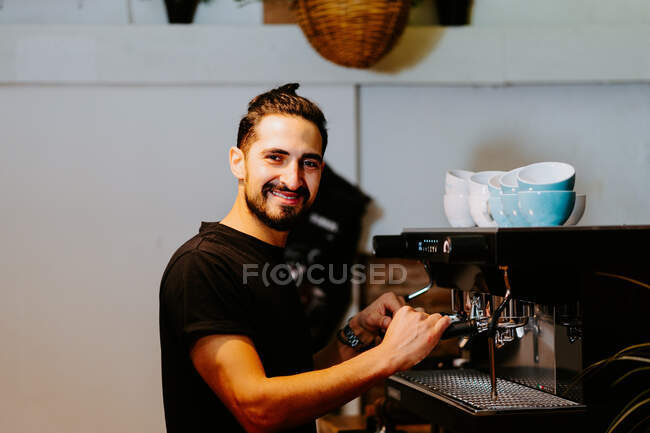Seitenansicht eines lächelnden männlichen Baristas, der Portafilter benutzt und Kaffee in einer modernen Kaffeemaschine zubereitet, während er im Café am Tresen steht und in die Kamera blickt — Stockfoto