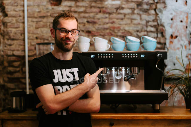 Lächelnder männlicher Barista mit Portafilter und Kaffeezubereitung in moderner Kaffeemaschine, während er im Café am Tresen steht und in die Kamera blickt — Stockfoto